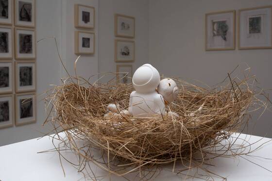 UStronaut und eine weitere Figur in einem Nest, im Hintergrund gerahmte Wandarbeiten.