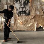 Man in a hat sweeping with a broom in front of the paper wall, a large black-and-white face above.
