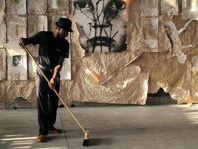Man in a hat sweeping with a broom in front of the paper wall, a large black-and-white face above.
