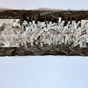 Lange, schmale Arbeit: Papierstreifen mit Schrift auf schwarzem, zerknittertem Papier, rote F&auml;den h&auml;ngen an den Enden.