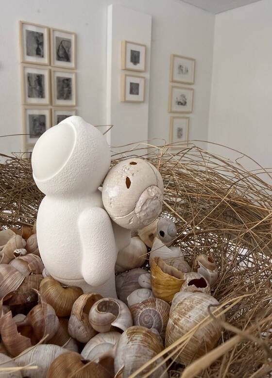 White figure in a nest of straw and snail shells, framed works in the background.