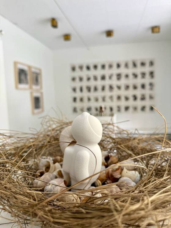UStronaut in a straw nest full of snail shells, the wall of small works in the background.