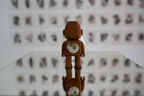 Rust-brown RAWbot from behind, with a wall of small paper works blurred in the background.