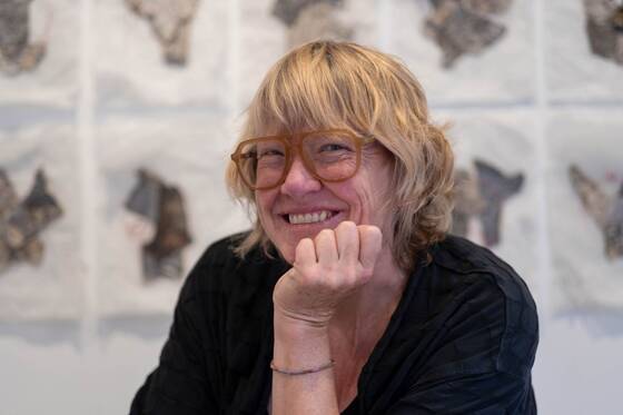 Smiling woman with glasses resting her chin on her hand, paper works hanging behind her.