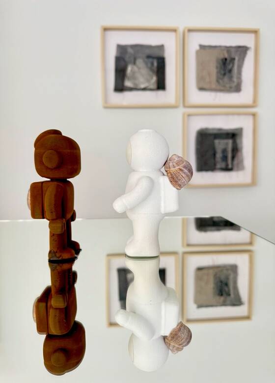 A RAWbot and a white UStronaut reflected in a mirrored surface in front of framed works.