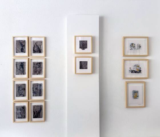 Small framed works in pale wooden frames on a white wall, arranged in three groups.