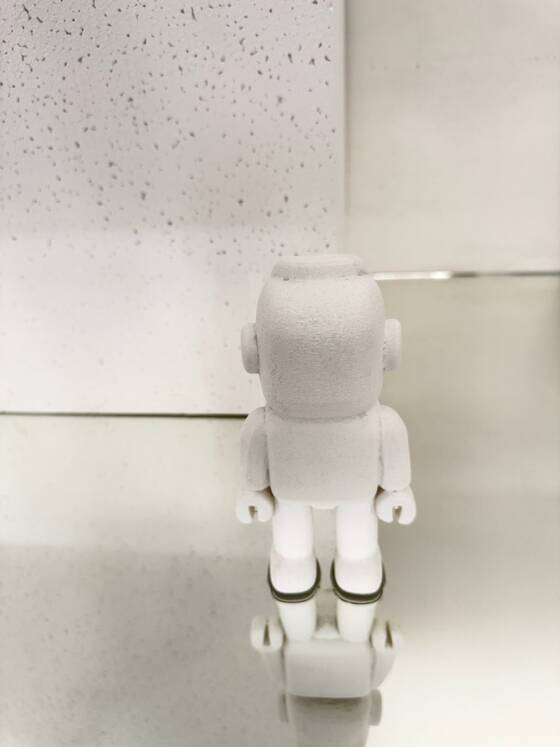 White UStronaut from behind on a mirrored surface in front of a light wall.