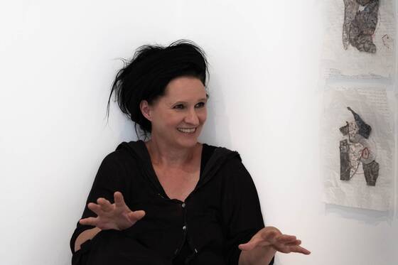 Dark-haired woman talking and gesturing, paper works on the wall beside her.